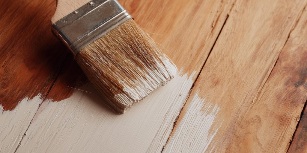 A paintbrush applying a base layer to a raw wood surface to prepare it for a final wood polish.
